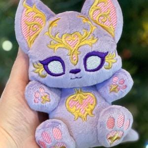 May include: A small, light purple plush toy shaped like a cat with embroidered gold and pink filigree patterns on its forehead, ears, and chest. It features large, dark purple-rimmed eyes with silver centers and pink paw pads on its feet.