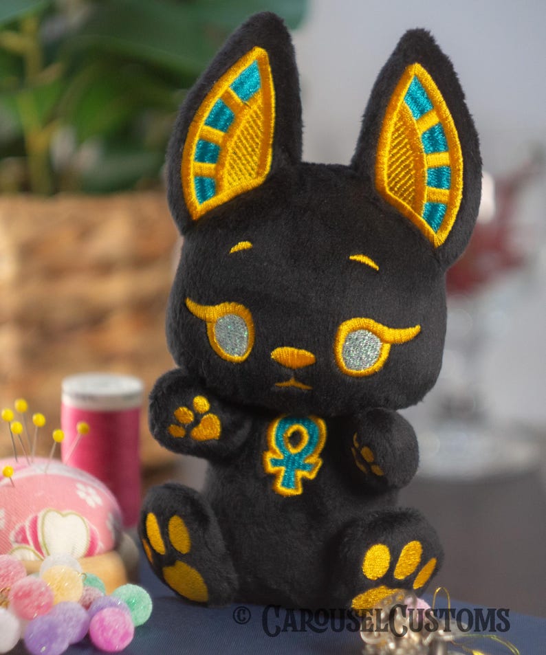 May include: Black plush toy with embroidered details. The toy is shaped like a cat with large ears and a golden Ankh symbol on its chest. The ears are outlined in gold and have a teal blue design. The eyes are sparkly silver.