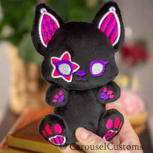 May include: Black plush toy cat with pink and purple ears, a pink star with a silver glitter eye on one eye, and a silver glitter eye on the other eye. The cat has pink paw pads.
