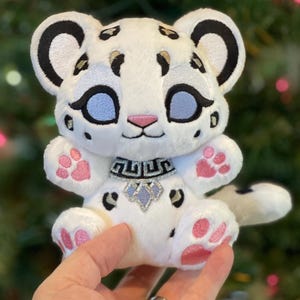 Pode incluir: Um peluche de leopardo-das-neves com pelo branco e manchas pretas, grandes olhos azul-claros, nariz cor-de-rosa e almofadas cor-de-rosa nas patas. A personagem usa uma coleira prateada e preta com um pendente geométrico e bordados detalhados.