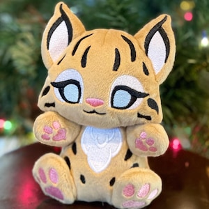 May include: A plush lynx toy with tan fur, black ear tufts, and black spots. It features large embroidered light blue eyes with black outlines, a small pink nose, and a white chest patch. The paws have embroidered pink paw pads on the feet.