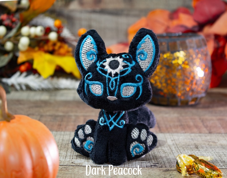 May include: Black plush toy with blue and silver embroidery, resembling a fox or cat with a single eye. The toy is sitting on a wooden surface with a pumpkin and a decorative candle holder in the background. The text "Dark Peacock" is visible below the toy.