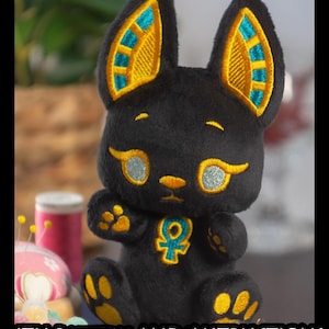 May include: Black plush toy cat with gold and teal embroidered ears, eyes, and paws. The toy has an embroidered ankh symbol on its chest. The toy is sitting on a table with other sewing supplies.