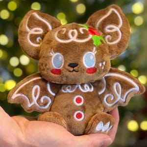 以下が含まれることがあります： 茶色のジンジャーブレッドクッキーのようなコウモリのぬいぐるみ。白いアイシングのディテールが施されています。コウモリは大きな耳、翼、青い目と赤い頬の笑顔の顔をしています。赤と緑のヒイラギの小枝で飾られています。