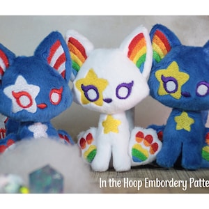 May include: Three plush toy cats with embroidered details. The cat on the left has a blue body with red and white stripes on its ears and a white star on its chest. The middle cat has a white body with rainbow stripes on its ears and a yellow star on its chest. The cat on the right has a blue body with rainbow stripes on its ears and a yellow star on its chest.  "In the Hoop Embroidery Pattern" is written below the cats.