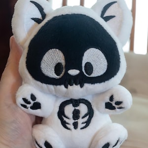May include: A white plush toy in the shape of a bear with a black skull-like face, paws, and chest emblem. The bear has large, embroidered eyes and a stitched mouth. The toy is soft and cuddly, with a whimsical design.