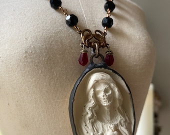 Puede incluir: Un collar de cuentas negras con un colgante de camafeo blanco que presenta una cara de calavera. El colgante está unido a una cadena dorada con dos cuentas rojas colgantes.