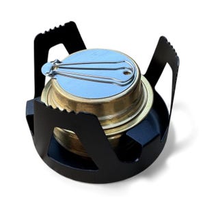 May include: A small camping stove featuring a brass fuel chamber and a silver lid. The stove is housed in a black metal frame with notched edges. Suitable for outdoor cooking and backpacking, it measures approximately 7 cm in diameter.