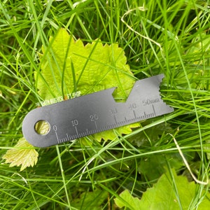 May include: A black metal multi-tool with a ruler marked in millimetres, up to 50mm. The tool has a hole, a notched edge, and various cutouts. The tool is lying on a green leaf and grass. The text "THE PATH BUSHCRAFT SHOP" is visible.