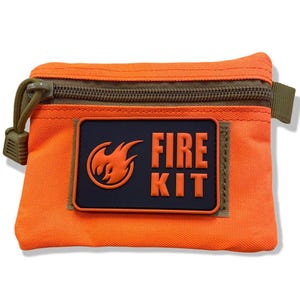 May include: An orange rectangular pouch with a dark green zip and pull tab. A black patch with the words "FIRE KIT" and a flame logo is centered on the front. The pouch is designed for outdoor survival.