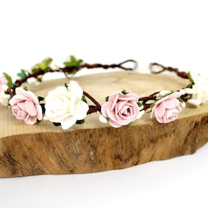 May include: A delicate floral crown with pink and white paper roses on a brown vine base. The crown is displayed on a wooden surface.