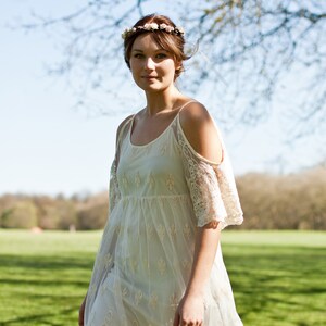 May include: A woman wearing a white lace dress with a floral crown. The dress has a cold shoulder design and is sheer. The woman is standing in a field with green grass and trees in the background.