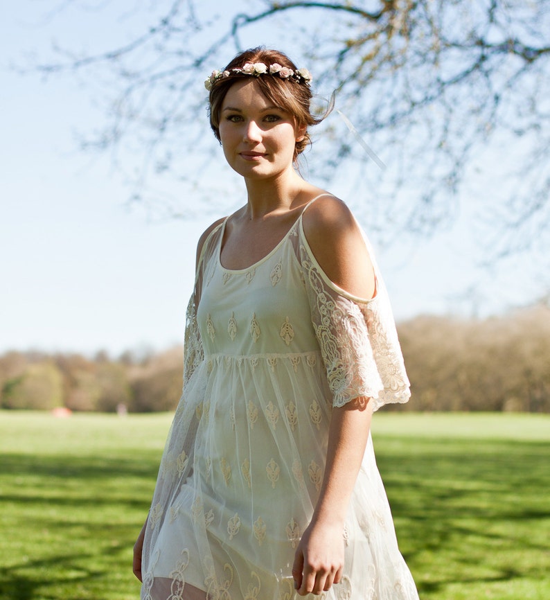 May include: A woman wearing a white lace dress with a floral crown. The dress has a cold shoulder design and is sheer. The woman is standing in a field with green grass and trees in the background.