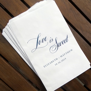 May include: White paper favor bags with the text "Love is Sweet" in a blue cursive font. The bags are stacked on top of each other and have the names "Elizabeth + Matthew" and the date "10.26.2019" printed below the main text.