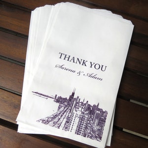 May include: A stack of white paper bags with a purple print of a city skyline and the text "THANK YOU Sirena & Adam".