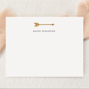 May include: A white card with a gold arrow pointing to the right and the text "KATIE STOCKTON" printed below the arrow.