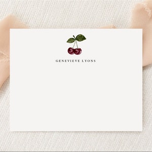 Puede incluir: Una tarjeta blanca con un diseño sencillo que presenta dos cerezas rojas con hojas verdes y el texto "Genevieve Lyons" impreso en negro.
