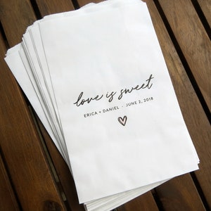 May include: A stack of white paper bags with the text "love is sweet" printed on the front. The bags also include the names "Erica + Daniel" and the date "June 2, 2018".