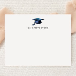 May include: A white card with a watercolor illustration of a graduation cap in shades of blue. The name "GENEVIEVE LYONS" is printed in black below the cap. The card is set against a soft, neutral background.