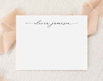 May include: A rectangular white stationery card featuring the name olivia jameson printed in a black, flowing, cursive script font centered at the top of the card. The card is positioned against a soft, light peach-colored fabric background.