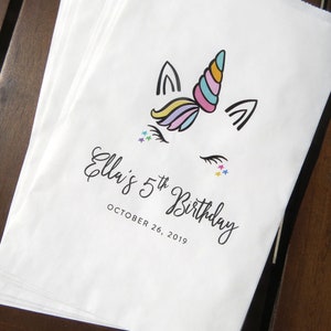 May include: White paper party favor bags with a unicorn illustration and the text "Ella's 5th Birthday" and "October 26, 2019".