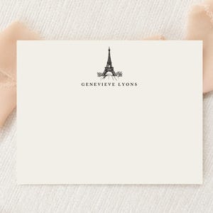 May include: A rectangular off-white stationery card featuring a centered black ink illustration of the Eiffel Tower. Below the tower, the name GENEVIEVE LYONS is printed in a clean, black serif font. The card has a smooth, matte finish.