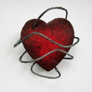 May include: A red heart-shaped object wrapped in barbed wire. The heart is made of paper mache and painted a deep red color. The barbed wire is made of metal and is wrapped around the heart in a random pattern.