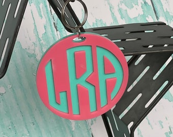 May include: A pink circle keychain with a teal monogram "LRA" on it. The keychain is attached to a silver split ring.