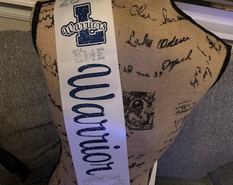 May include: White satin sash with blue glitter lettering that reads "2020 Senior Warriors". The sash has a blue and white logo with the letter "L" and the word "Warriors" on it.