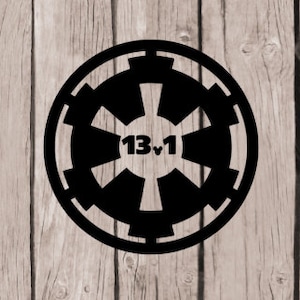 May include: Black and white Imperial symbol with the text "13♥1" in the center. The symbol is on a wood background.