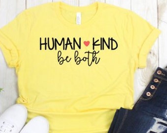 May include: Yellow short-sleeved t-shirt with the text "HUMAN KIND be both" in black lettering with a red heart between the words "HUMAN" and "KIND". The shirt is laying on a white wooden surface with a pair of white sneakers and blue jeans.