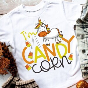 May include: White t-shirt with a cartoon unicorn and the text "I'm a Candy Corn". The shirt is surrounded by fall-themed decorations, including a denim jacket, pumpkins, and fall leaves.