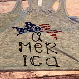K&ouml;nnte beinhalten: Ein graues Tanktop mit einem glitzernden rot-wei&szlig;-blauen amerikanischen Flaggen-Design und dem Wort "America" in Schwarz geschrieben mit einem kleinen roten Herzen im "e".