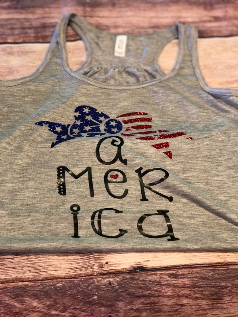 K&ouml;nnte beinhalten: Ein graues Tanktop mit einem glitzernden rot-wei&szlig;-blauen amerikanischen Flaggen-Design und dem Wort "America" in Schwarz geschrieben mit einem kleinen roten Herzen im "e".