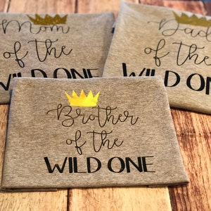 May include: Three gray t-shirts with black text and gold crowns. The shirts say "Mom of the Wild One", "Dad of the Wild One", and "Brother of the Wild One".