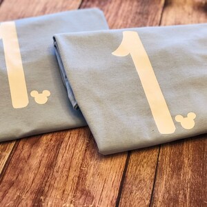 May include: Two light blue t-shirts with a white number "1" and a white Mickey Mouse silhouette on each.