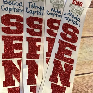 May include: Four white sashes with red glitter letters spelling out "SENI" and the words "EHS Cheer" and the names "Becca Captain", "Tehva Captain", "Aldai Captain", and "Jaeda Captain" written in blue ink.