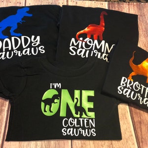 May include: Four black t-shirts with dinosaur silhouettes and text. The shirts read "Daddy Sauraus", "Mommy Sauraus", "Brother Sauraus", and "I'm One Colten Sauraus".