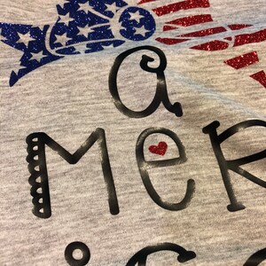 K&ouml;nnte beinhalten: Ein graues T-Shirt mit einem glitzernden rot-wei&szlig;-blauen amerikanischen Flaggen-Design. Der Text "A merica" ist in Schwarz mit einem kleinen roten Herzen im "e" geschrieben.