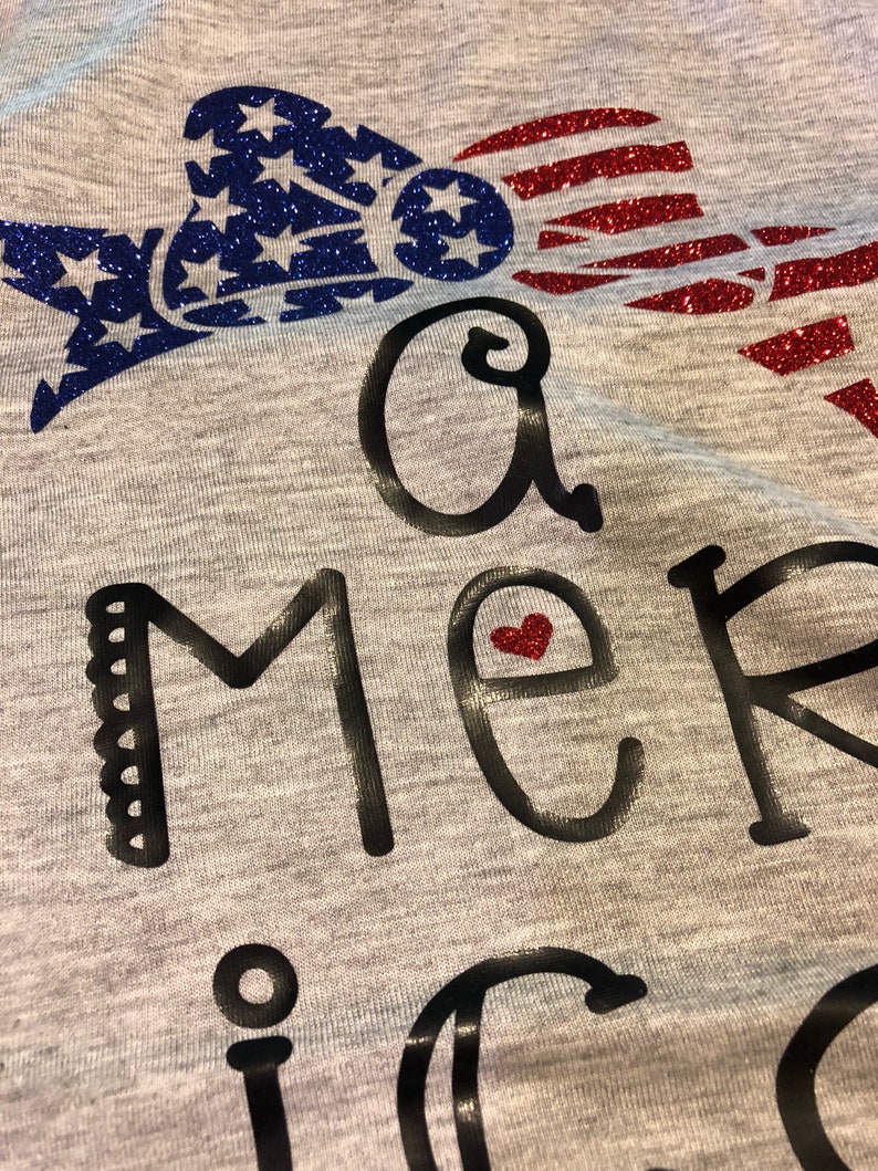 K&ouml;nnte beinhalten: Ein graues T-Shirt mit einem glitzernden rot-wei&szlig;-blauen amerikanischen Flaggen-Design. Der Text "A merica" ist in Schwarz mit einem kleinen roten Herzen im "e" geschrieben.