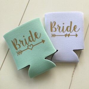 Pode incluir: Dois isoladores de bebidas, um branco e outro verde claro, com a palavra "Bride" impressa em letras douradas e uma flecha com um cora&ccedil;&atilde;o.
