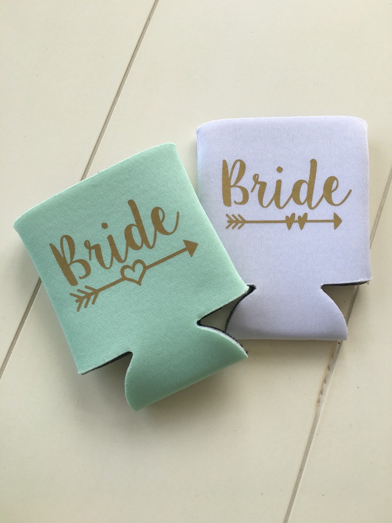 Pode incluir: Dois isoladores de bebidas, um branco e outro verde claro, com a palavra "Bride" impressa em letras douradas e uma flecha com um cora&ccedil;&atilde;o.