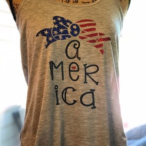 K&ouml;nnte beinhalten: Ein graues Tanktop mit einem roten, wei&szlig;en und blauen Glitzerdesign eines Bogens und dem Wort "America" mit einem roten Herzen.