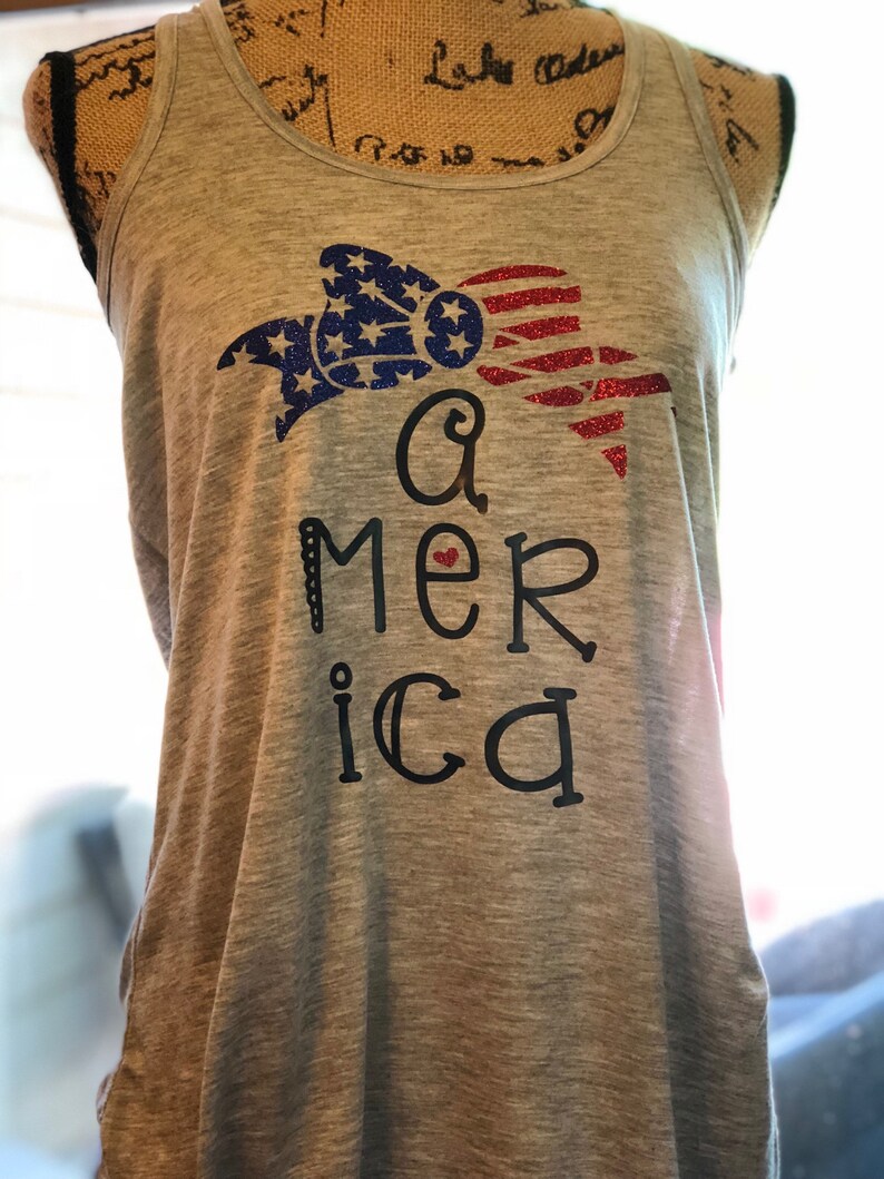 K&ouml;nnte beinhalten: Ein graues Tanktop mit einem roten, wei&szlig;en und blauen Glitzerdesign eines Bogens und dem Wort "America" mit einem roten Herzen.