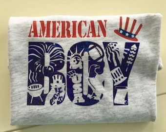 Puede incluir: Una camiseta gris con el texto "AMERICAN BOY" en rojo y azul. Las letras están formadas por imágenes de iconos estadounidenses, como fuegos artificiales, la Estatua de la Libertad, una pelota de béisbol y una parrilla.