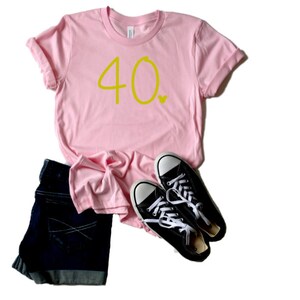 May include: A pink t-shirt with the number 40 in yellow lettering. The shirt is folded and laid flat on a white background with a pair of black sneakers.