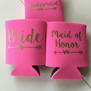 Pode incluir: Tr&ecirc;s refrigeradores de latas de espuma rosa com letras douradas. Os refrigeradores dizem "Bride", "Maid of Honor" e "Bridesmaid".