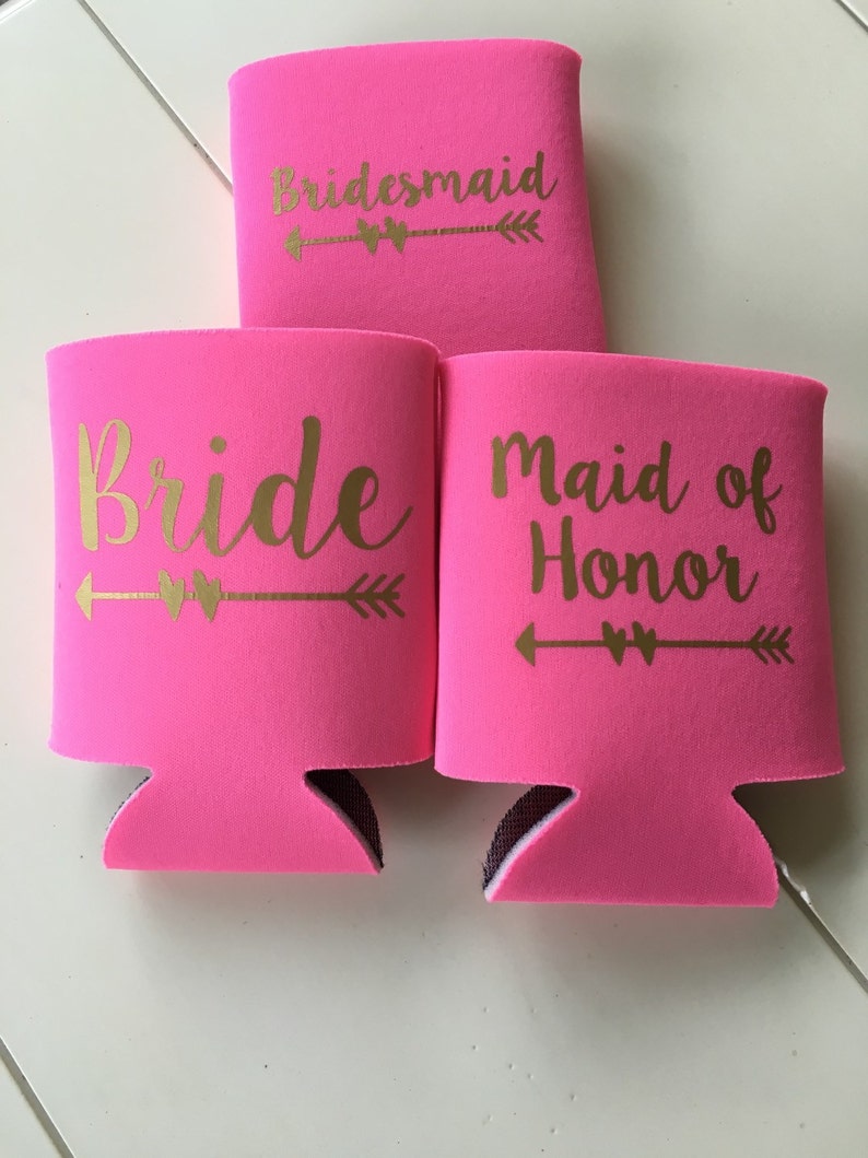 Pode incluir: Tr&ecirc;s refrigeradores de latas de espuma rosa com letras douradas. Os refrigeradores dizem "Bride", "Maid of Honor" e "Bridesmaid".