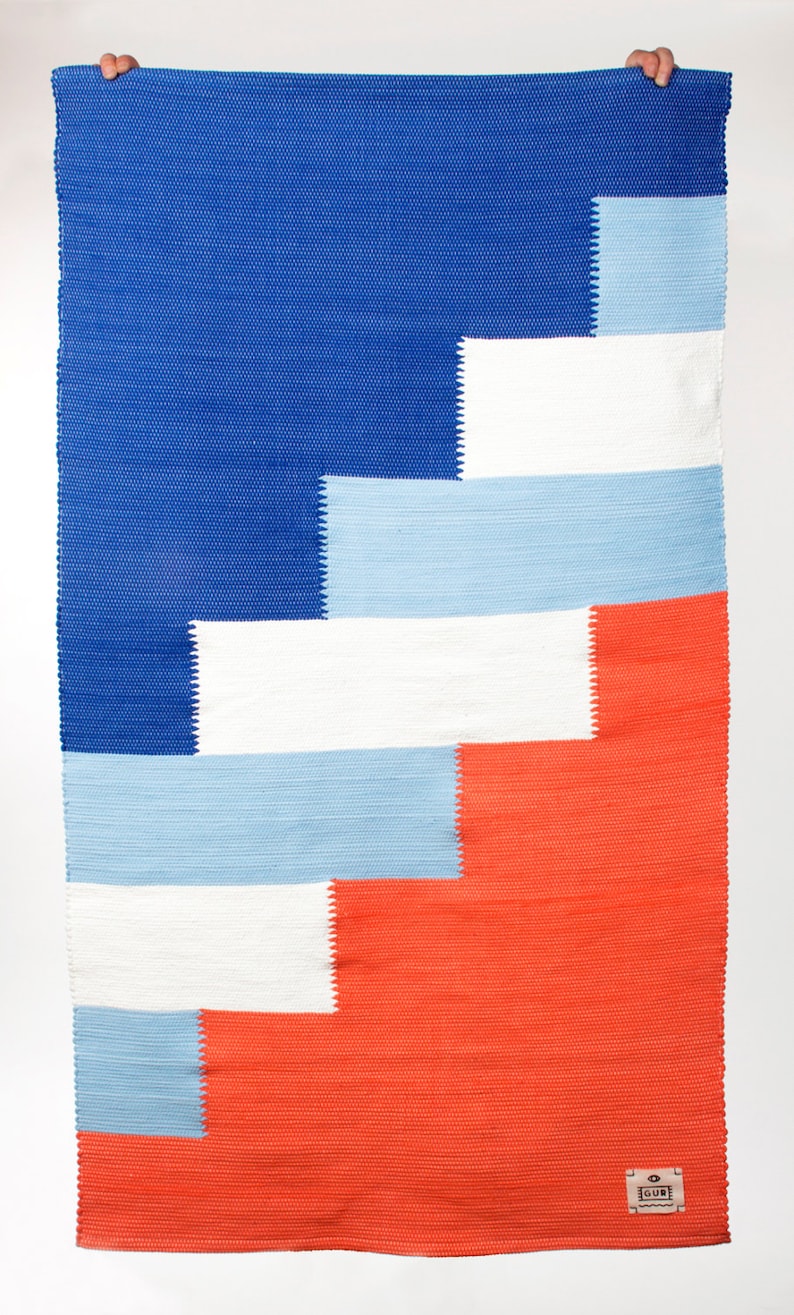 Op de afbeelding: Een geweven tapijt met een geometrisch patroon in tinten blauw, wit en oranje. Het tapijt heeft een label met de tekst "GUB" in de rechterbenedenhoek.