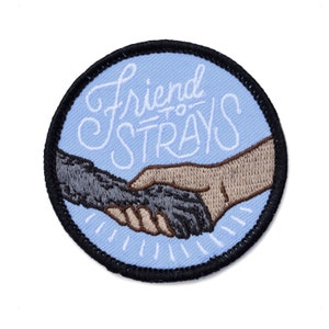 Op de afbeelding: Ronde patch met de tekst "Friend to Strays" in witte letters op een lichtblauwe achtergrond. De patch toont een hondenpoot die een hand schudt met een menselijke hand, beide in bruin- en grijstinten, met een zwarte rand.